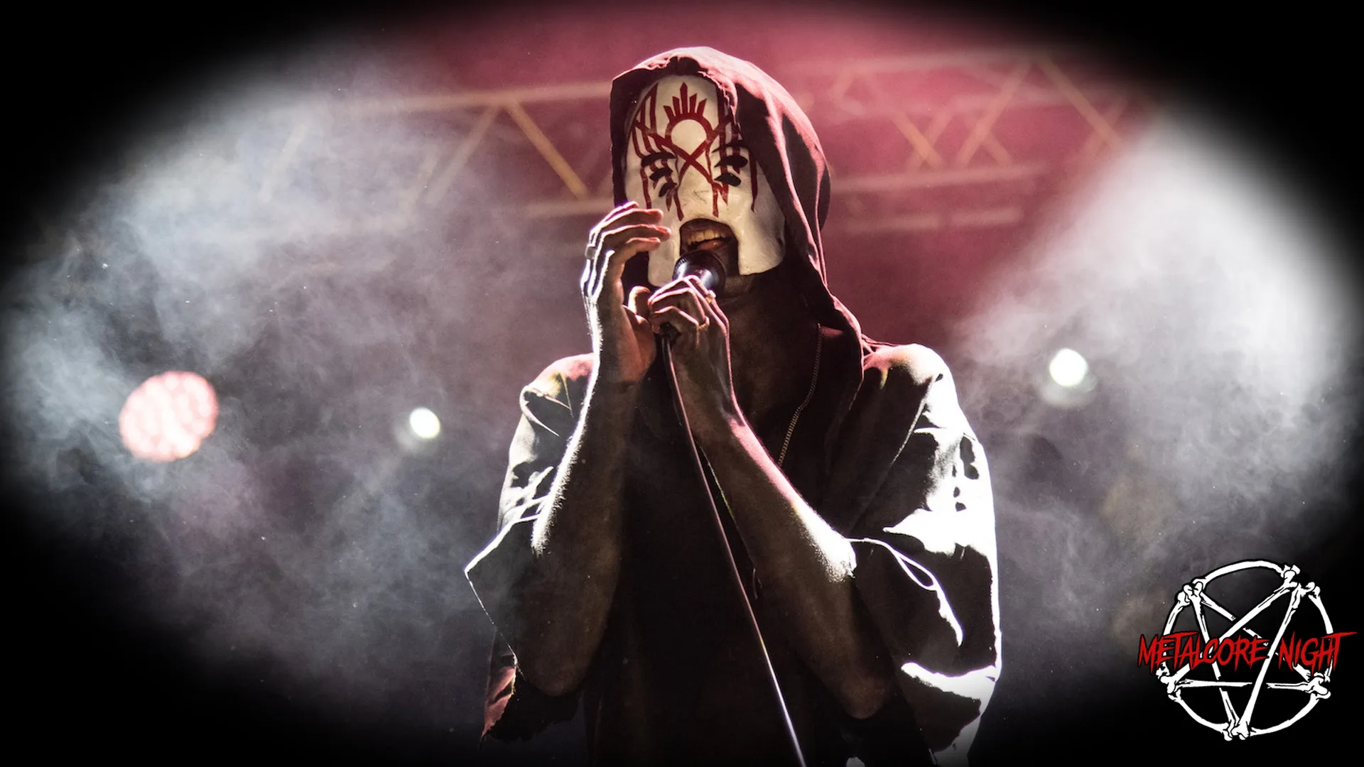 This image portrays a dramatic stage scene featuring a musician from the band Sleep Token, taken during a live performance. The band is played regularly at Metalcore Night events. The band member is centrally framed and shrouded in a mysterious aura. They wear a distinct mask with a red, intricate emblem resembling a sigil, adding a ritualistic element to their appearance. A hooded garment cloaks their figure, merging with the dark backdrop of the stage. The setting is low-lit, with the exception of spotlights that cast a soft glow on the performer, enhancing the intensity and focus on them. The musician's stance is one of deep engagement, with their hands cupping a microphone close to the mask's mouth opening, suggesting they are in the midst of a passionate vocal performance. The atmosphere is further electrified by a misty haze that envelopes the scene, catching the stage lights and creating a dreamlike quality. This adds to the enigmatic and otherworldly vibe synonymous with Sleep Token's image. The bottom right corner features a graphic logo for Metalcore Night, comprising an inverted pentagram and circular saw blade, reinforcing the edgy and hardcore theme of the event.
